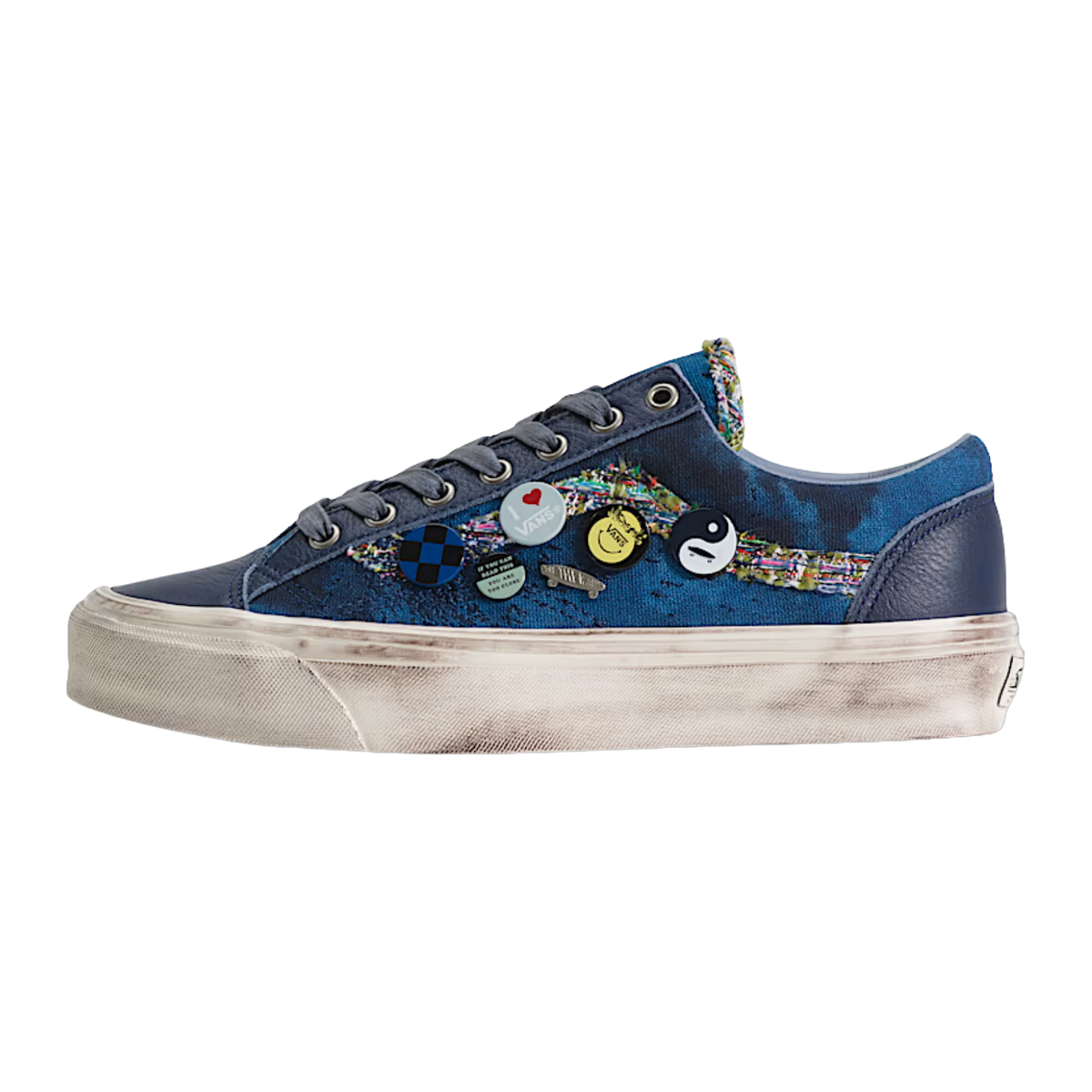 A low-top sneaker in navy blue canvas with a colorful abstract pattern on the side, decorated with several small circular pins, featuring a distressed off-white sole.